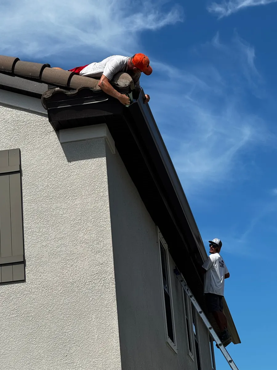 Two installers fitting a dark brown seamless gutter along the tile-roof edge of a two-story stucco home in Starkey Ranch, Odessa FL
