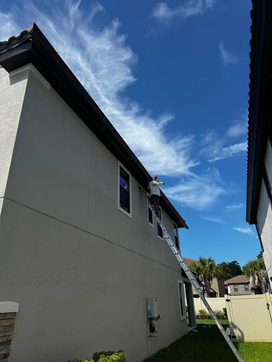 Installer on a ladder beside a long run of dark brown seamless gutters on a two-story stucco home in Starkey Ranch, Odessa FL