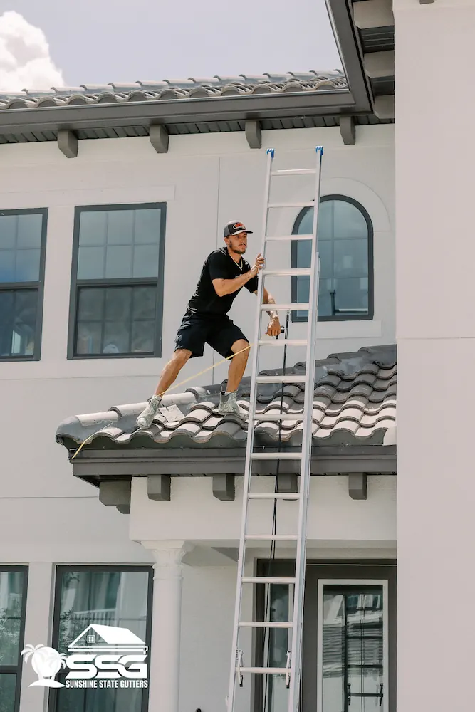 Installer safely accessing a tile-roof home using a ladder to prepare for a 7-inch gutter installation, as part of Sunshine State Gutters’ professional tile-roof service.